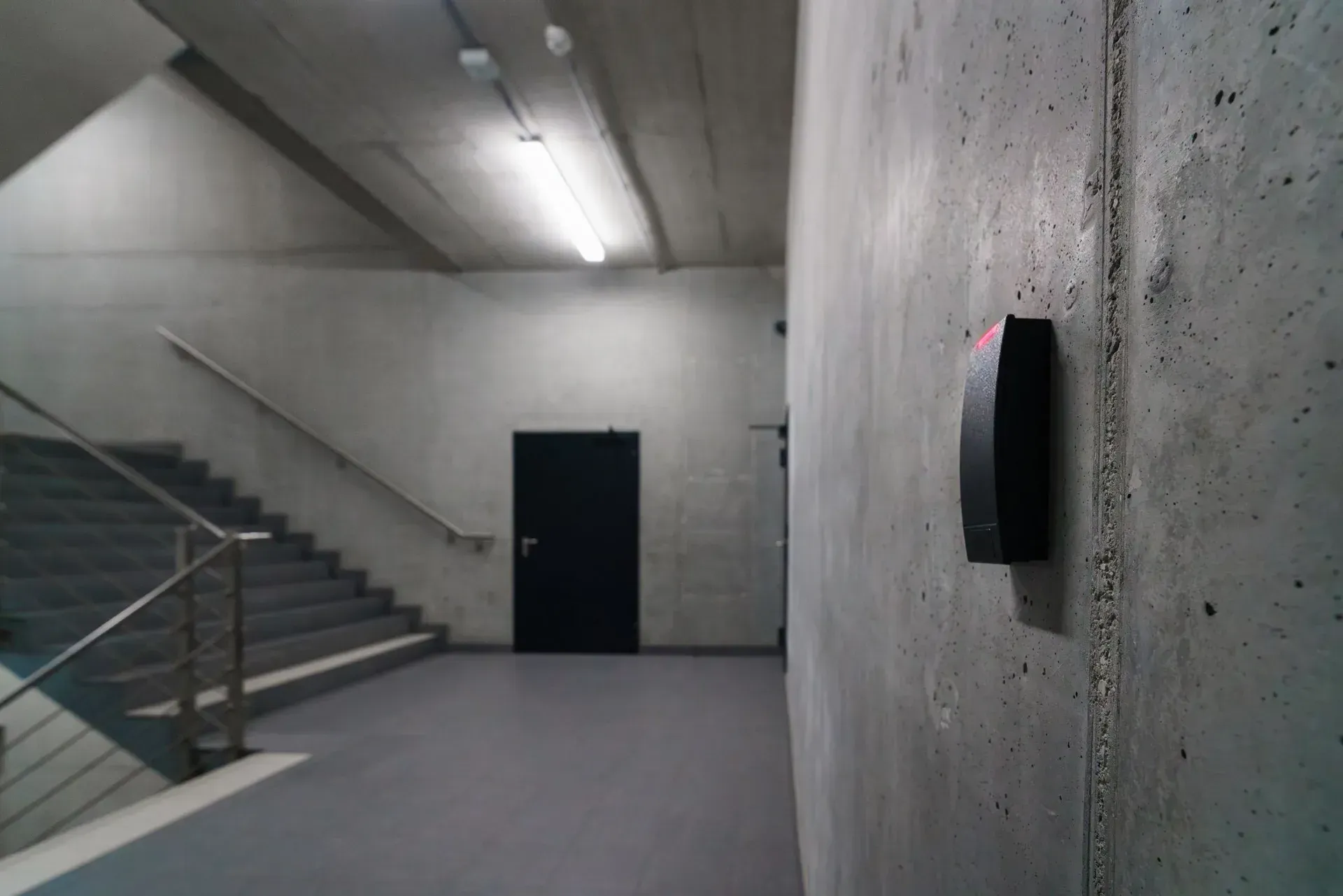 A black proximity reader on a concrete wall, with a corridor leading to a secured door behind it