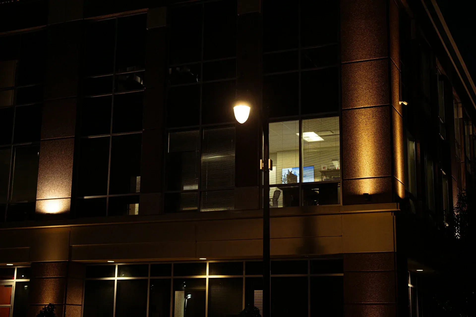 An office building at night with one window lit beside a street lamp.