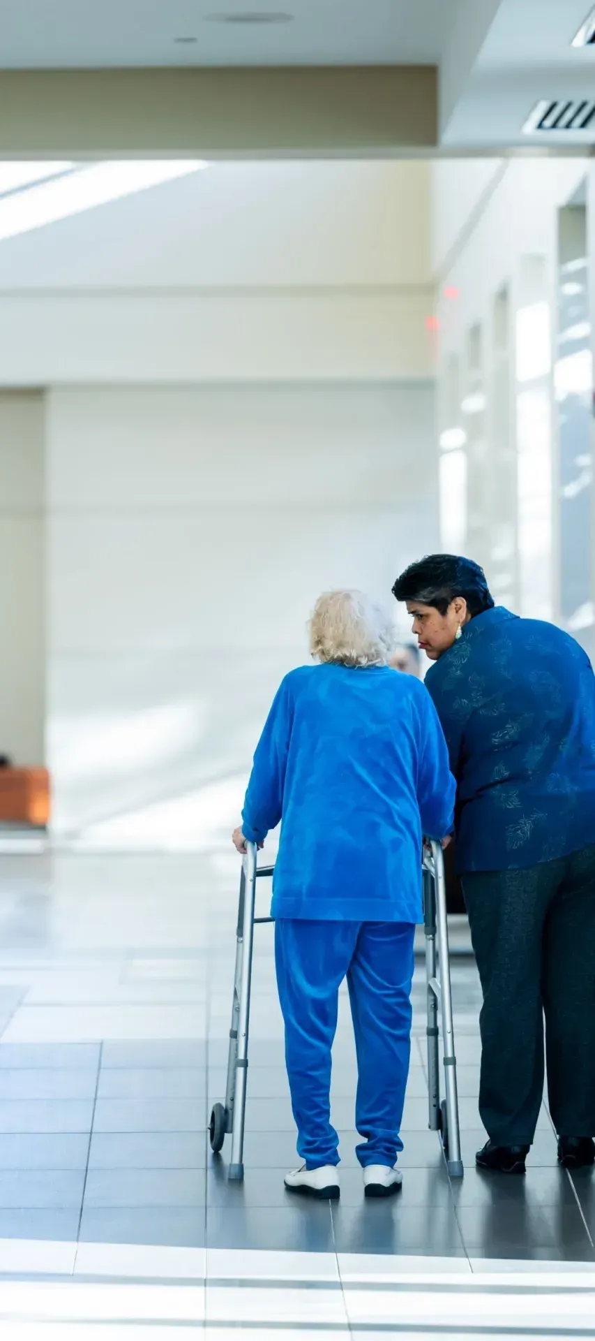 Nurse helping an older woman walk with a frame along a hospital corridor