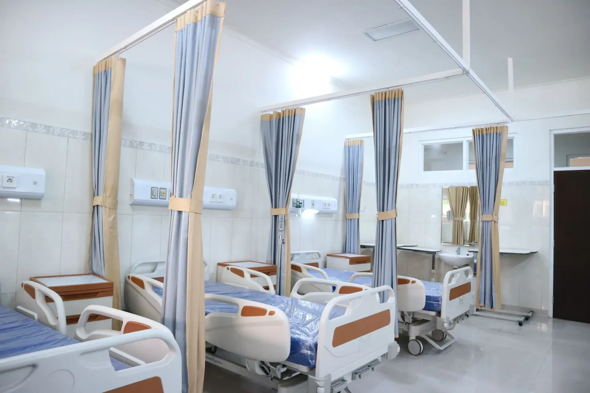 A bright hospital ward: beds, privacy curtains on ceiling tracks and a door to the next room.