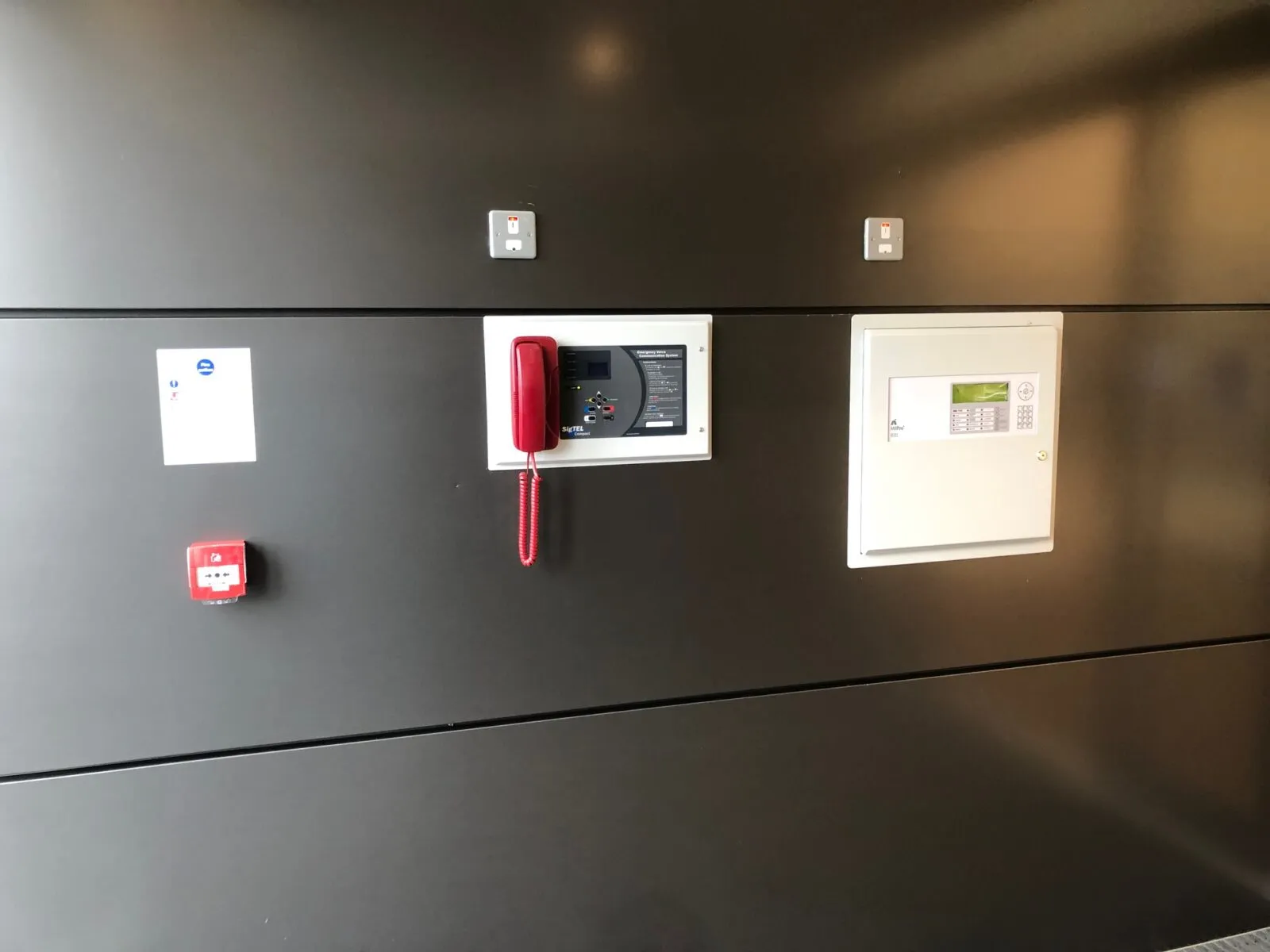 A fire alarm panel and a disabled refuge handset on a dark wall