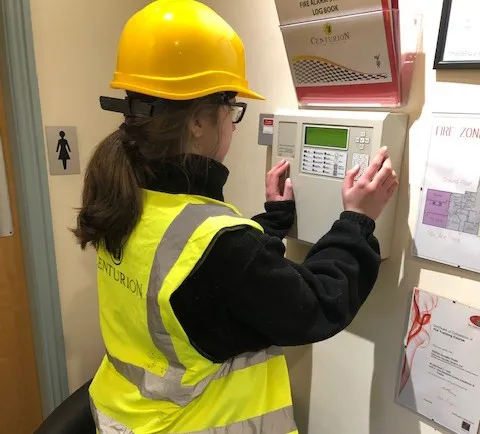 An apprentice in a yellow hard hat and high-visibility vest works on a fire alarm panel