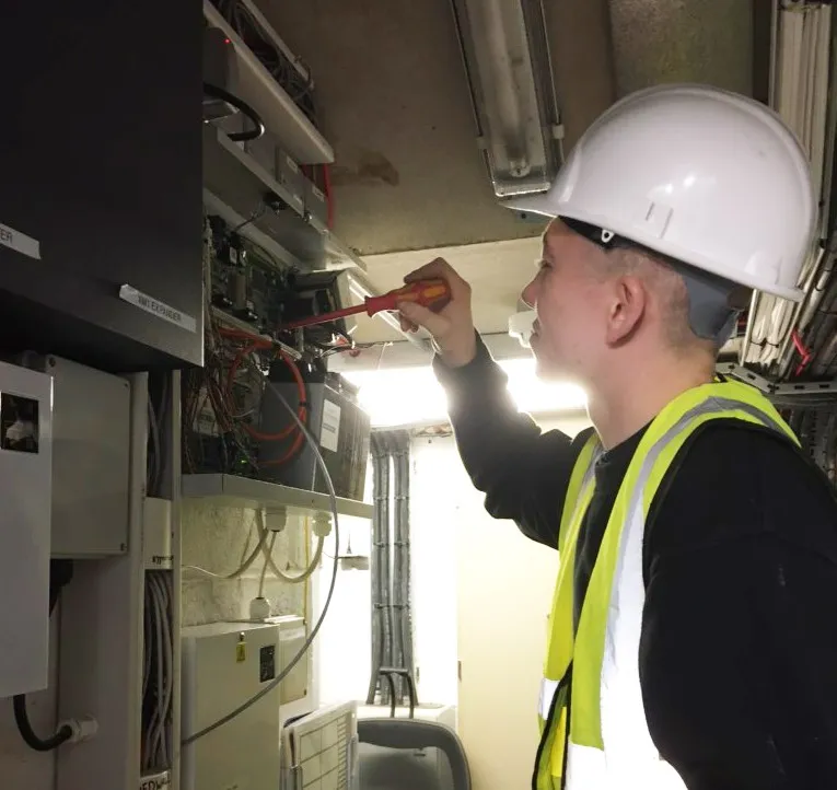 An apprentice in a white hard hat works inside an alarm panel with a screwdriver