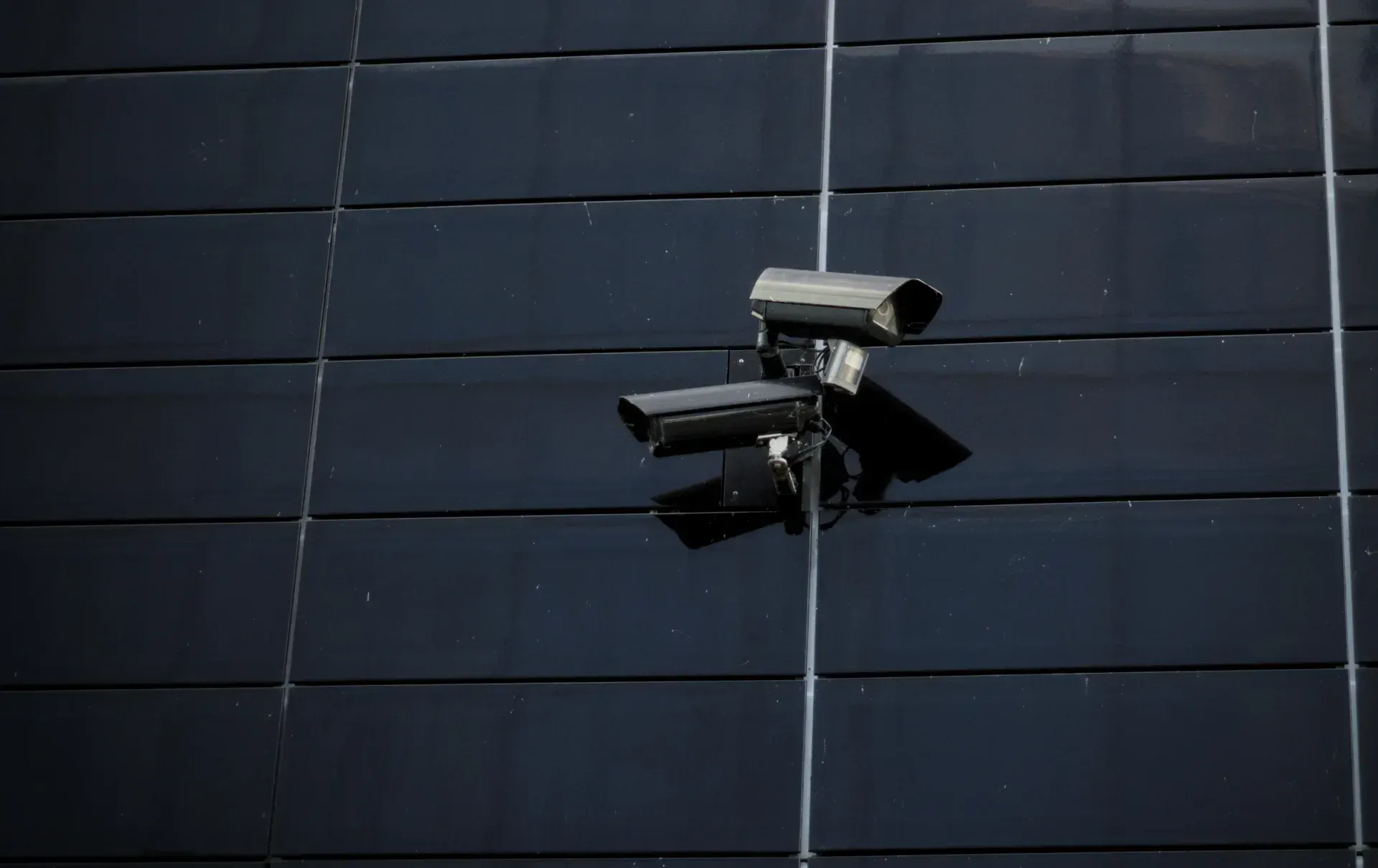 Two CCTV cameras in weatherproof housings on one bracket, on the dark cladding of a commercial building