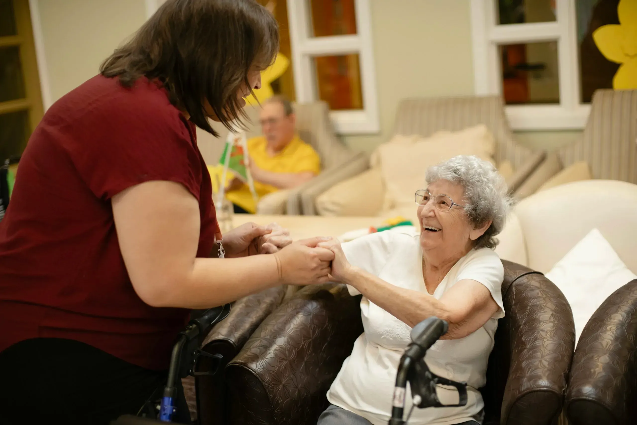 A carer holding the hands of a smiling resident in a care home lounge