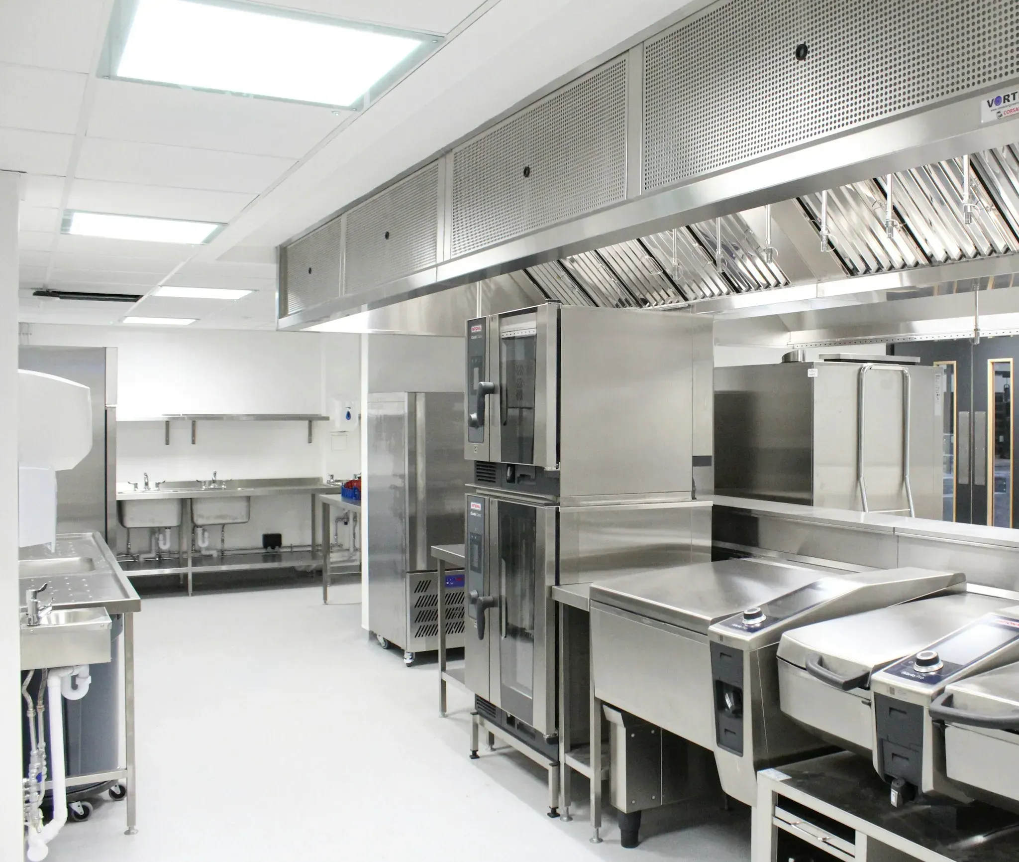 A bright commercial kitchen under a long stainless steel extraction canopy