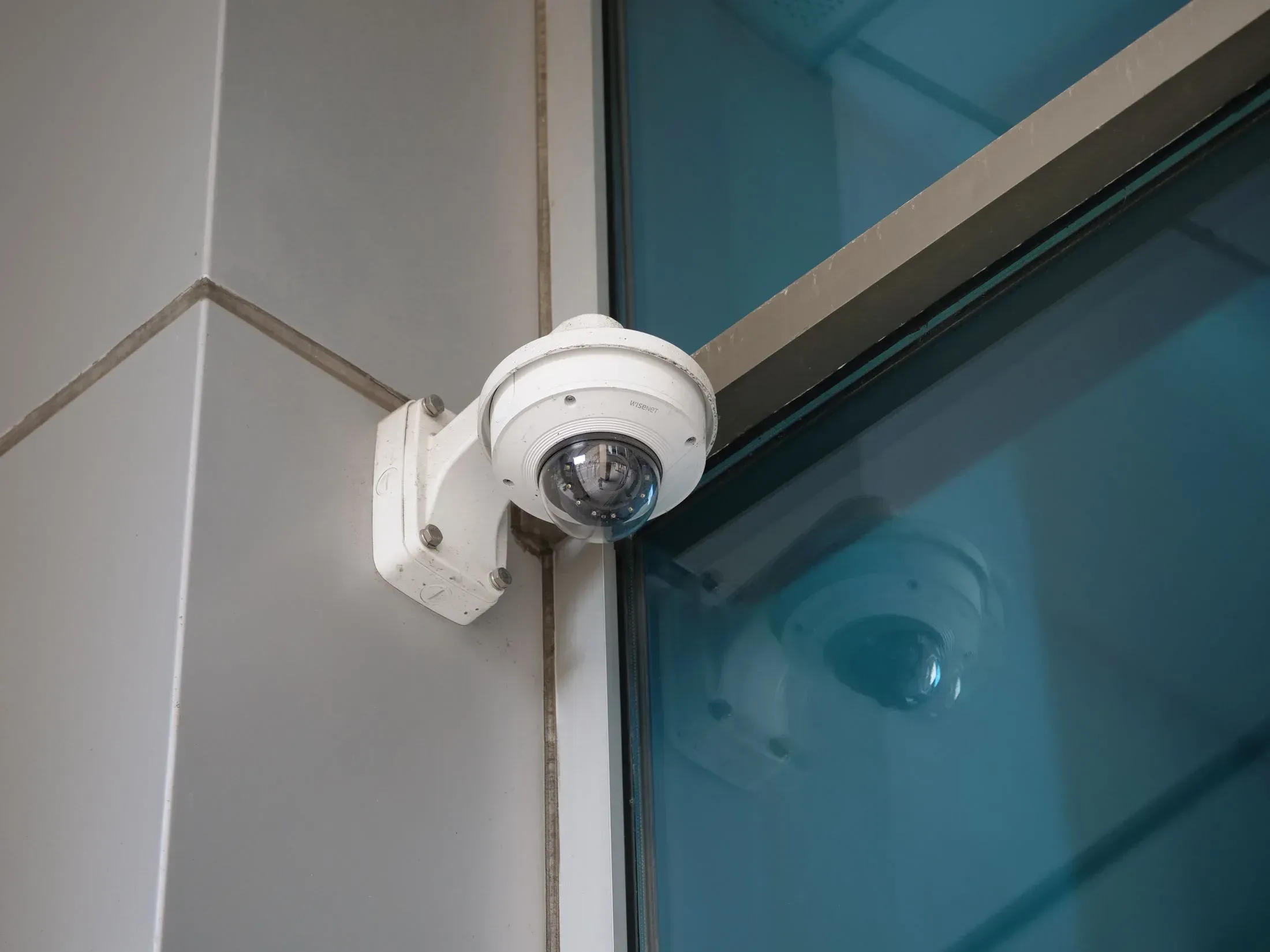 A white dome CCTV camera on a wall bracket beside blue glazing