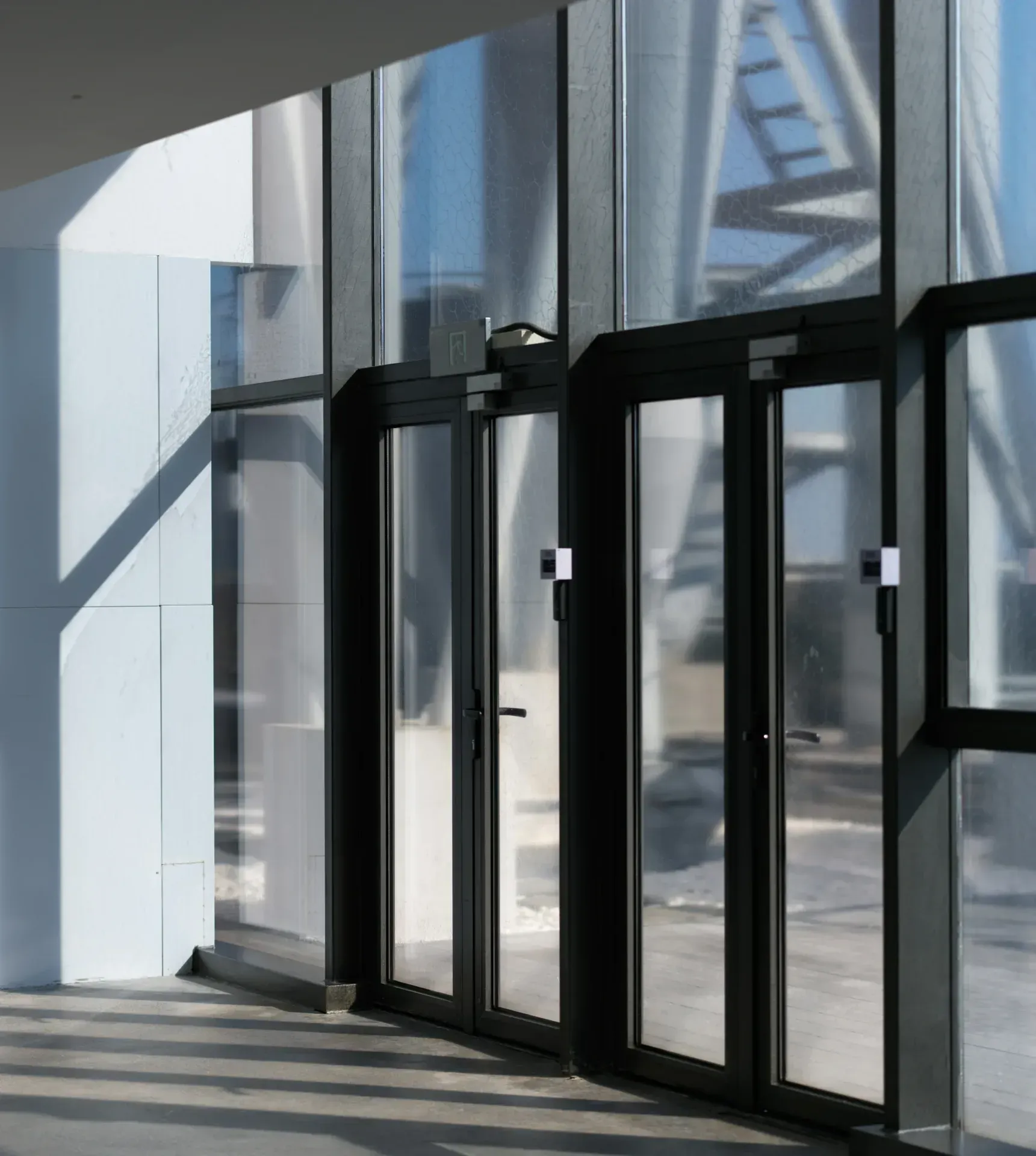 A row of glass entrance doors in raking sunlight, each with a slim access control reader on its frame.