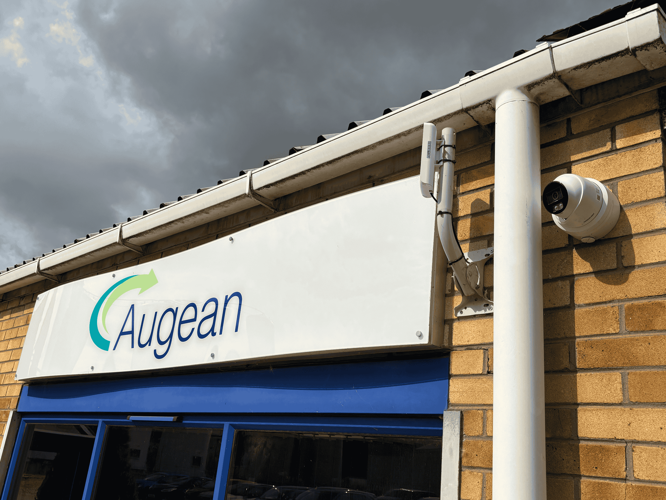 A fixed turret CCTV camera mounted on the exterior brick wall of a building beneath a white sign with the logo “Augean.