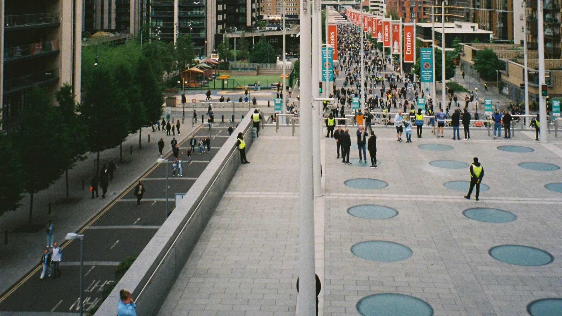 Stewards in high-visibility vests watch fans walk up Olympic Way towards Wembley Stadium on a match day.