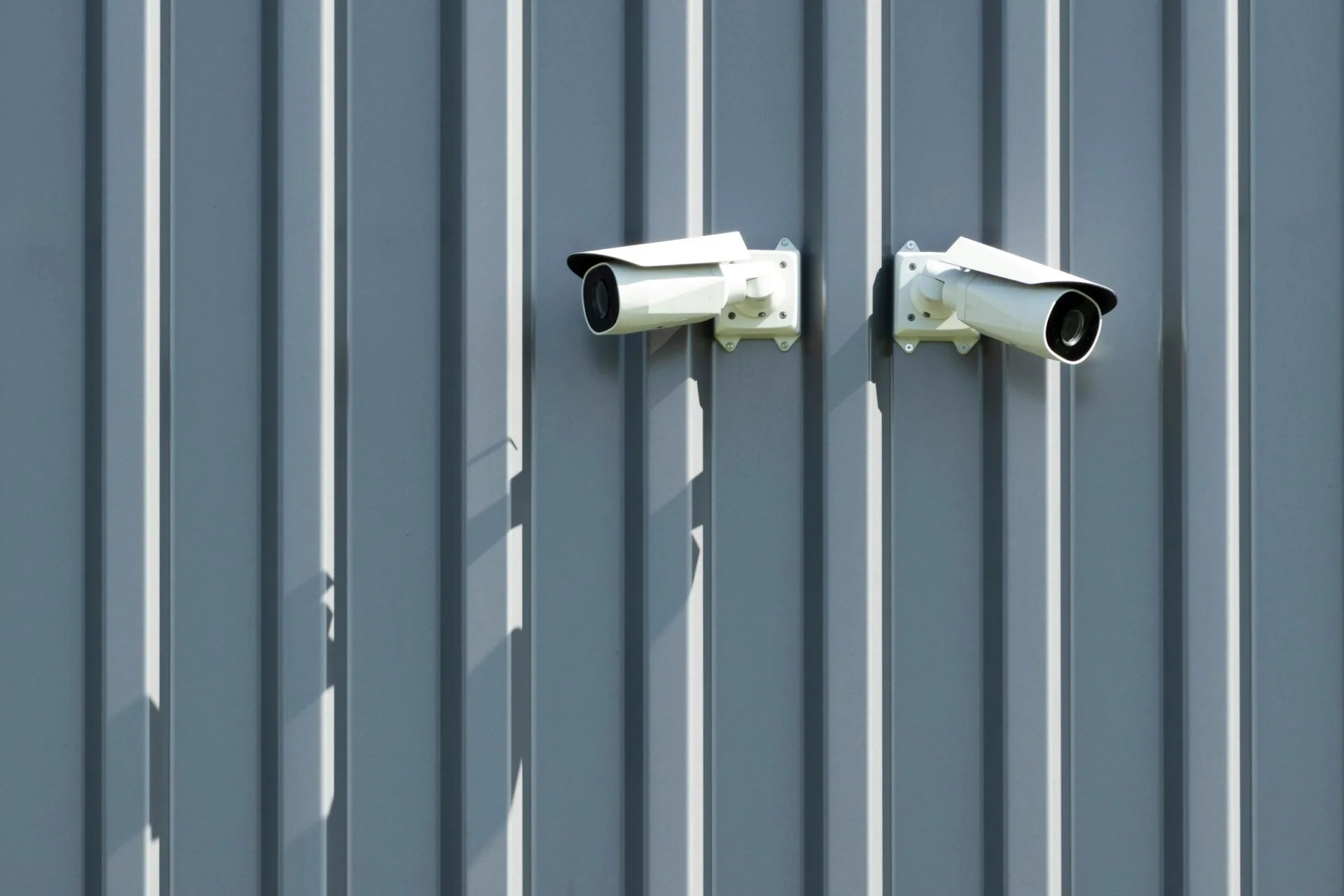 Two white bullet CCTV cameras mounted on blue-grey steel cladding in low sunlight