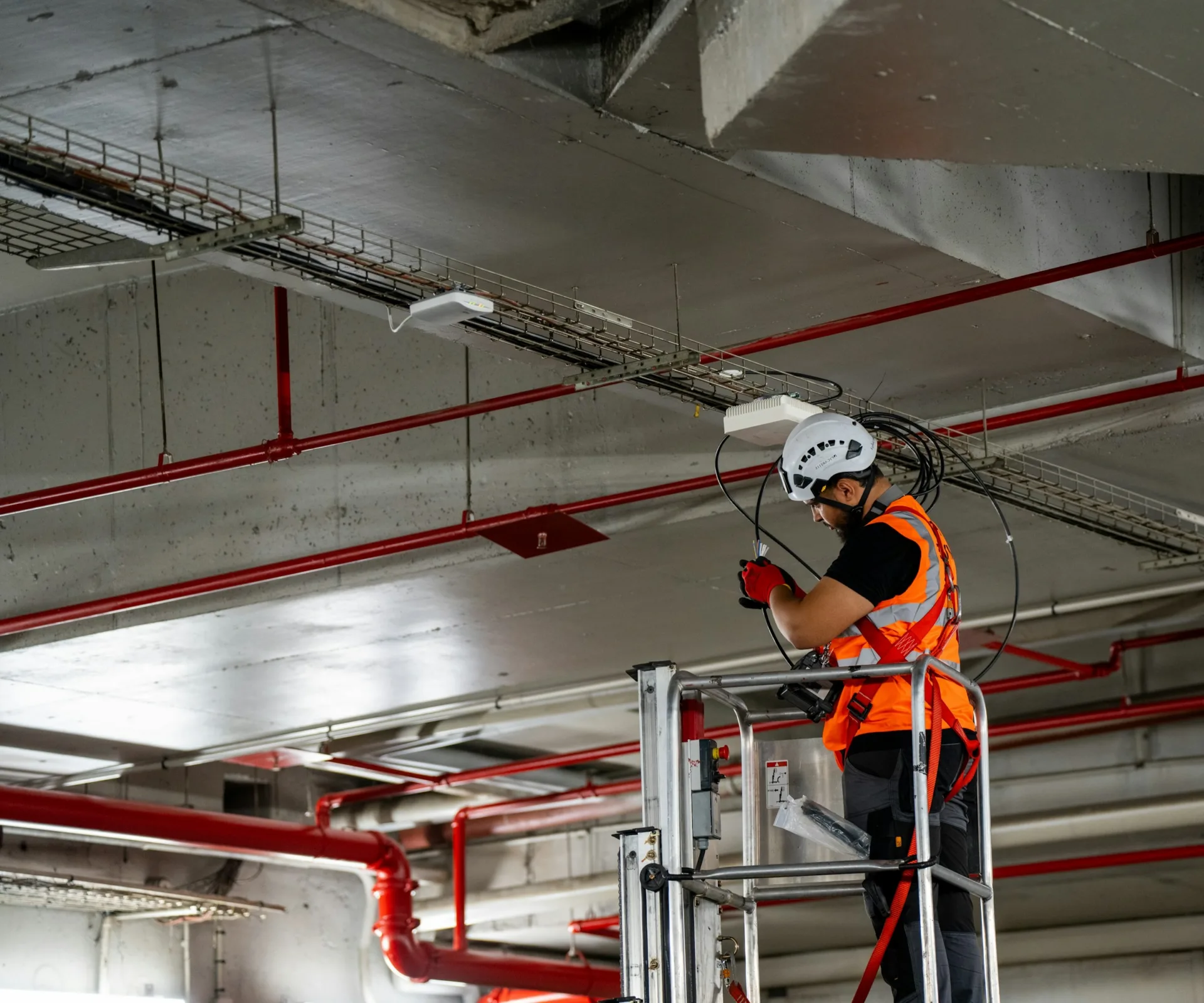 An engineer in a helmet and hi-vis on an access platform, working on ceiling-mounted equipment and cabling
