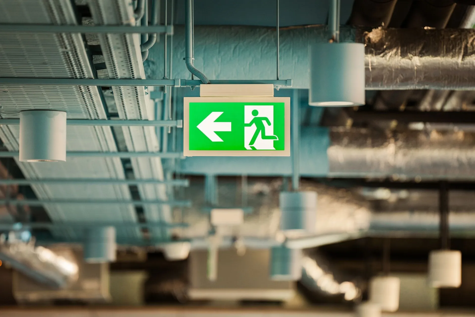 Green running-man fire exit sign hanging below exposed services in a modern commercial ceiling