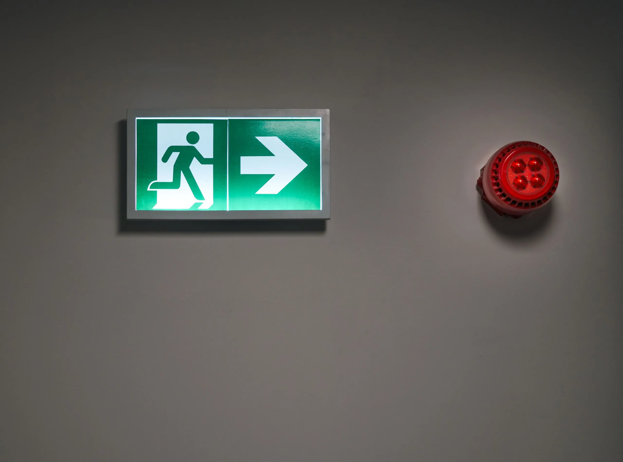 Lit exit sign beside a red sounder beacon on a grey wall