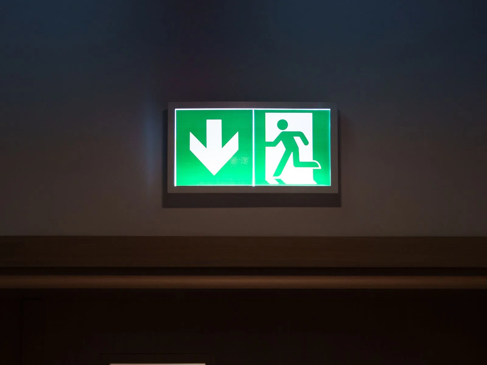 Illuminated green exit sign with a downward arrow above a closed door