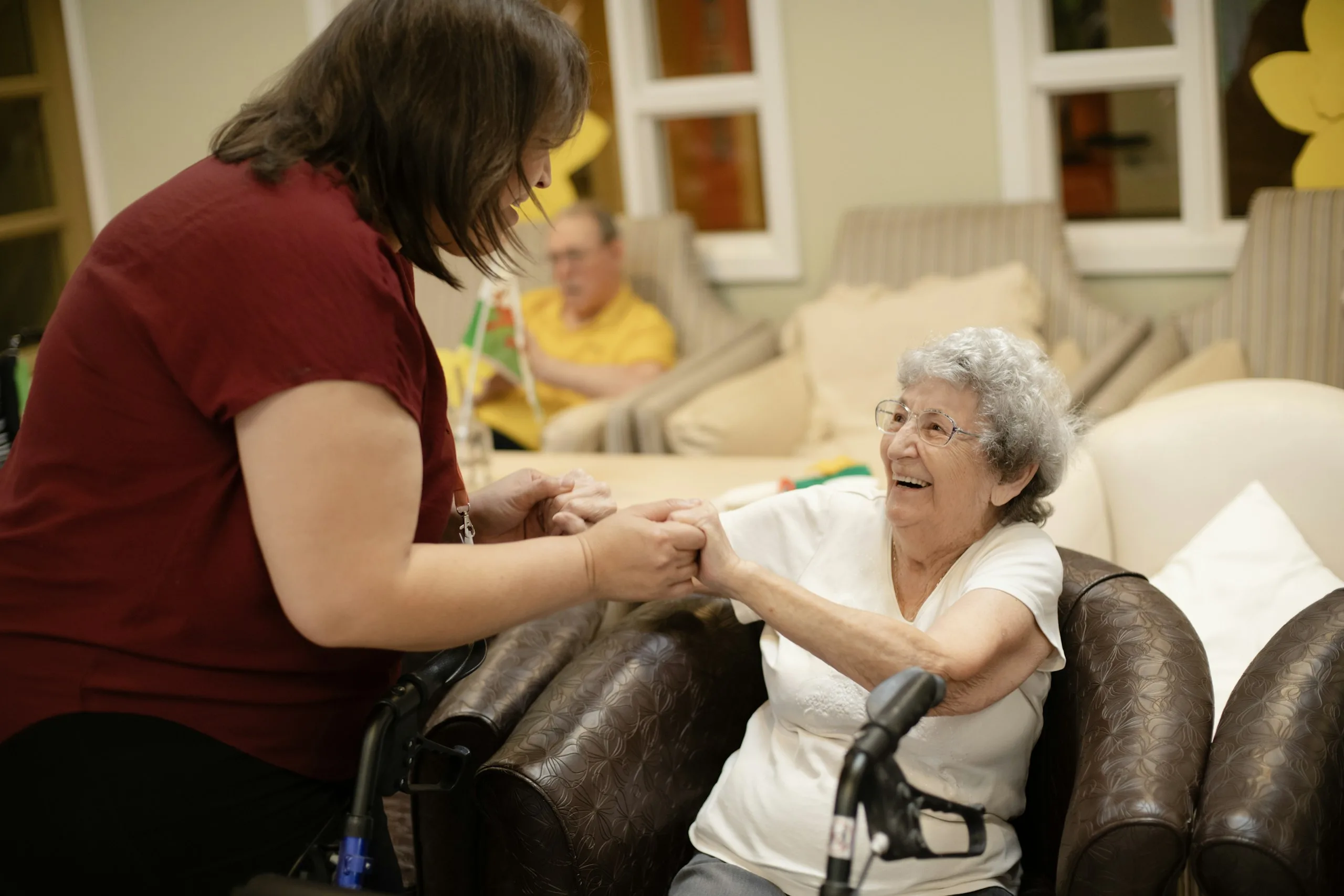 A carer holding the hands of a smiling resident in a care home lounge