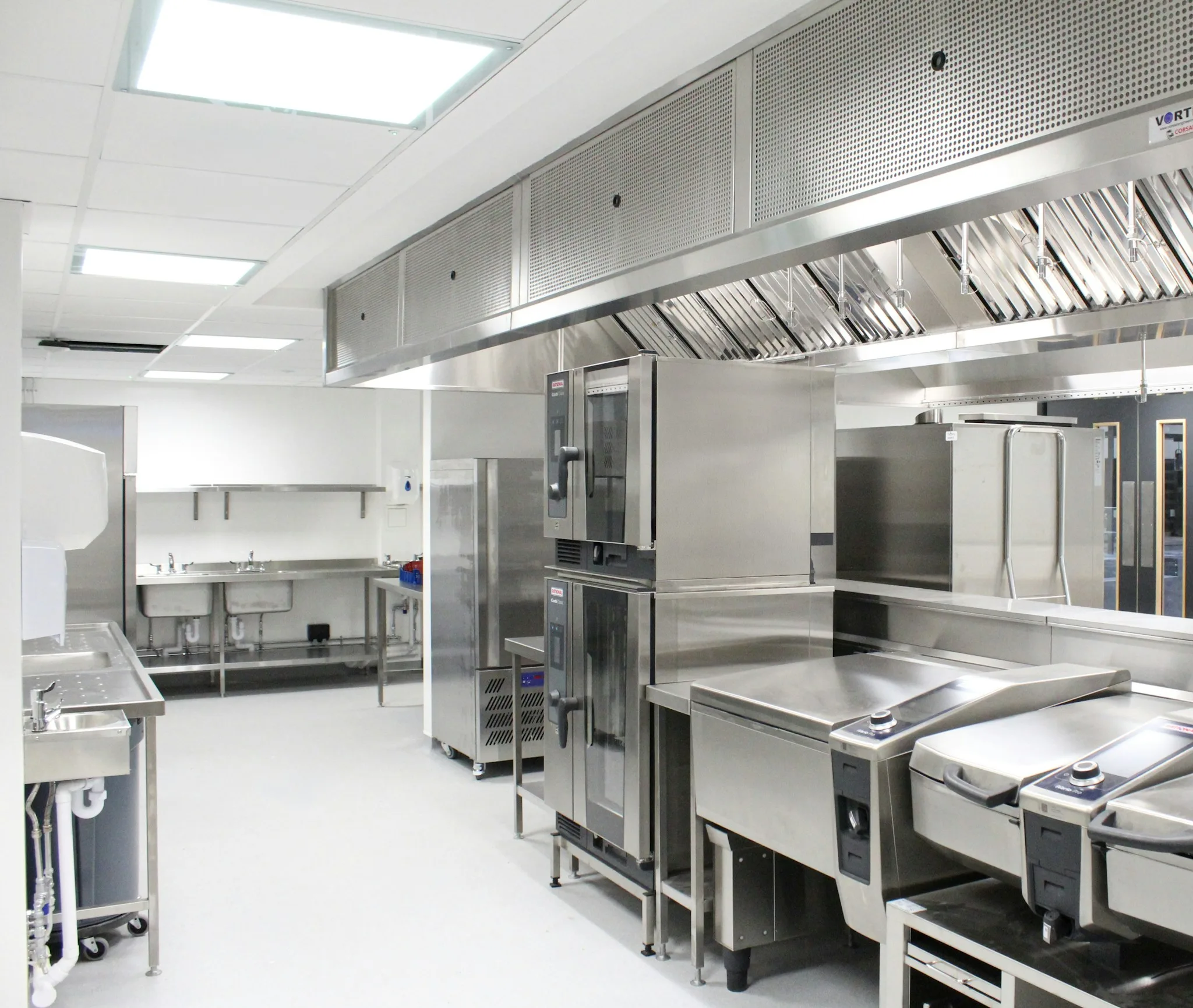 A bright commercial kitchen under a long stainless steel extraction canopy