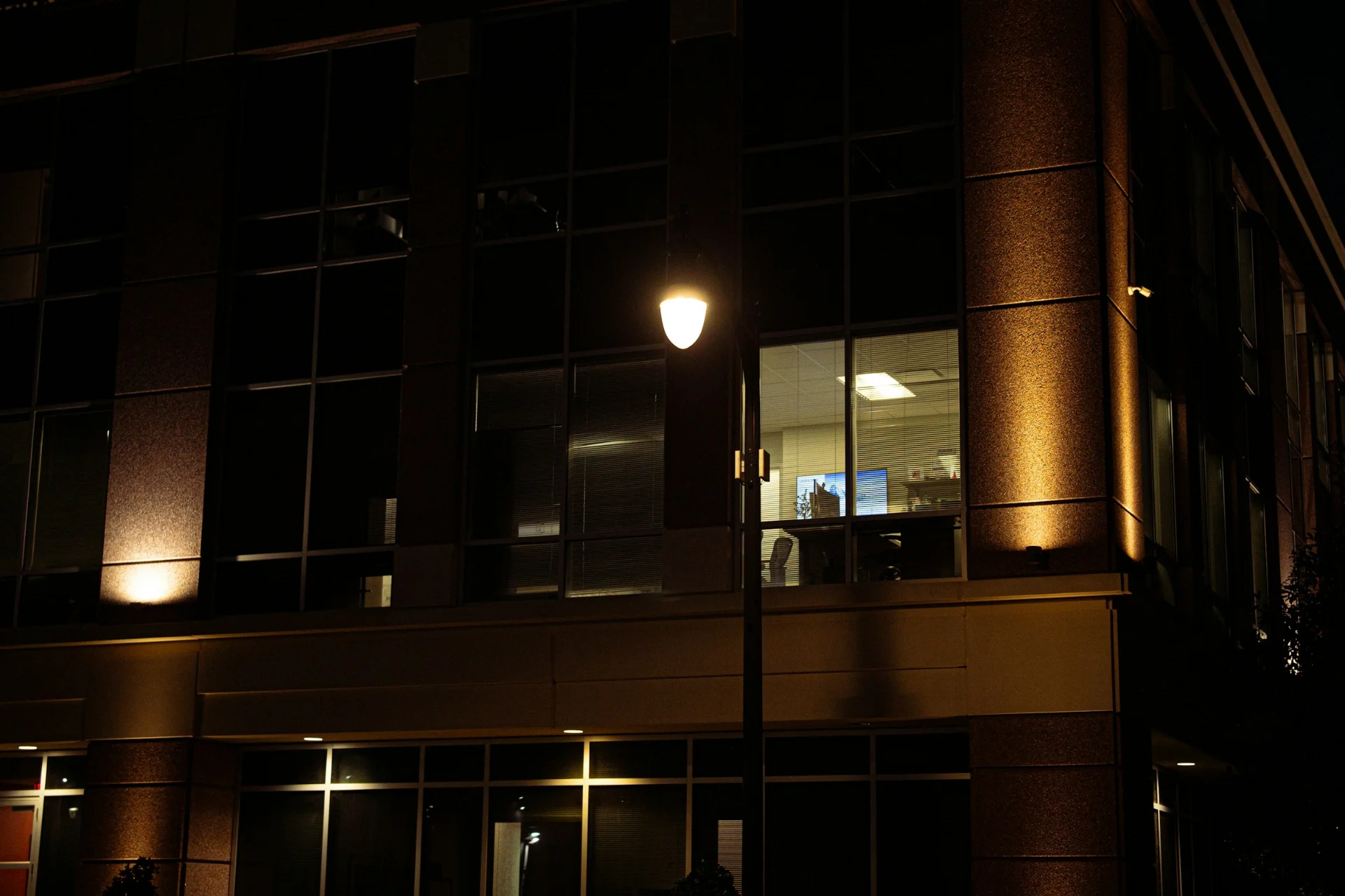 An office building at night with one window lit beside a street lamp.